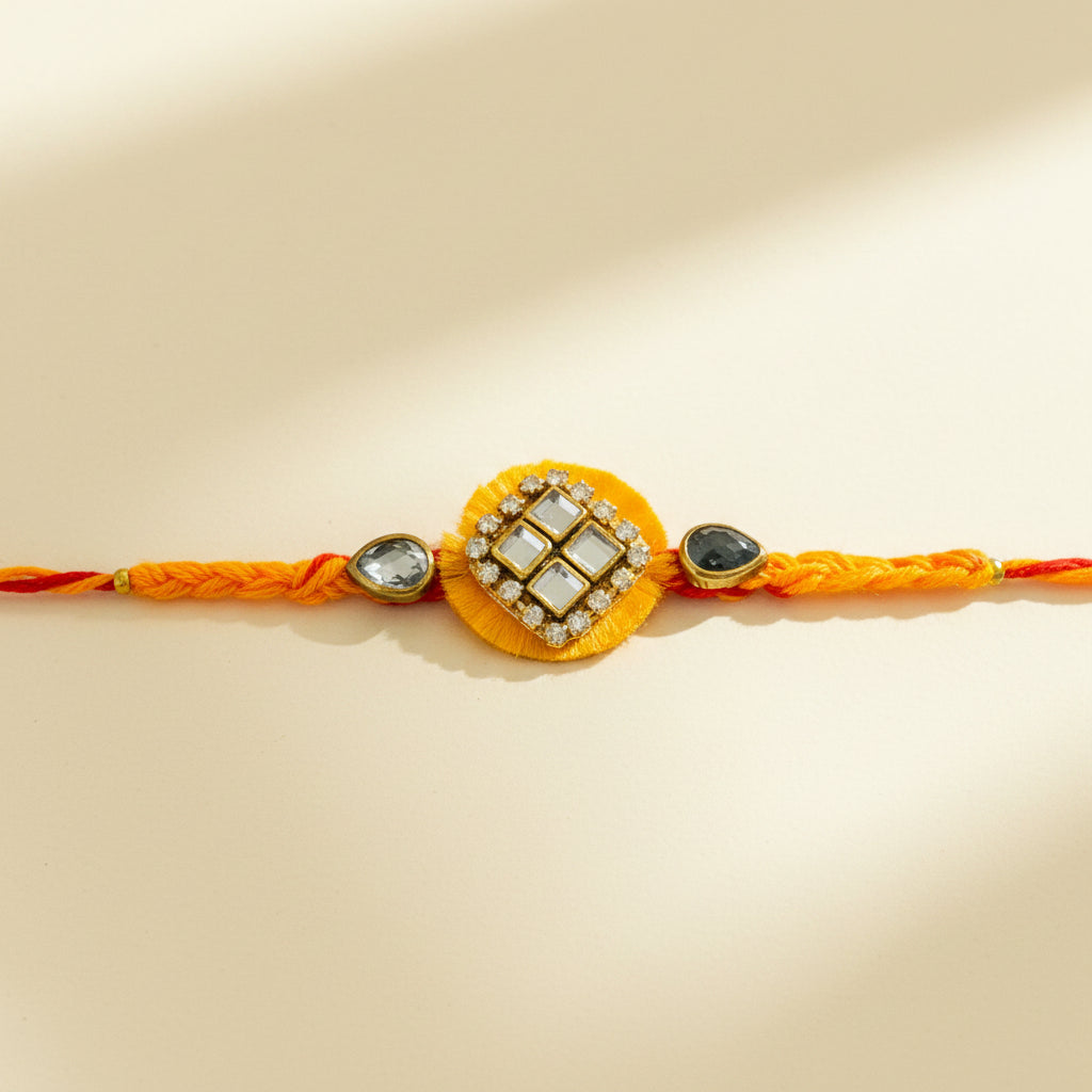 A traditional Indian Rakhi bracelet laid flat on a white flower-shaped paper prop over a woven straw mat. The bracelet features a bright orange and red braided cord with a central yellow circular ornament, a gold-toned square frame set with white stones, and small decorative beads on either side.