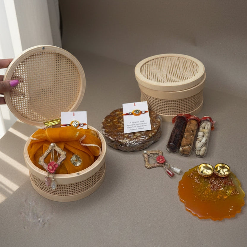 A collage showing a handmade Rakhi gift set in a round woven bamboo basket. The set includes a red and gold thread Rakhi on a white card, a folded orange fabric pouch with a decorative charm, a small wrapped item, three small gift tubes, a decorative metal piece, and a gold glittery orange resin piece.