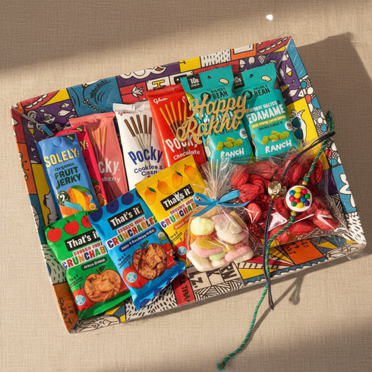 A colorful gift basket filled with assorted packaged snacks and sweets, arranged on a beige fabric background with decorative green leaves. Visible snack brands include Soley, Pocky, Happy Rakhe, That's It, and Ranch.