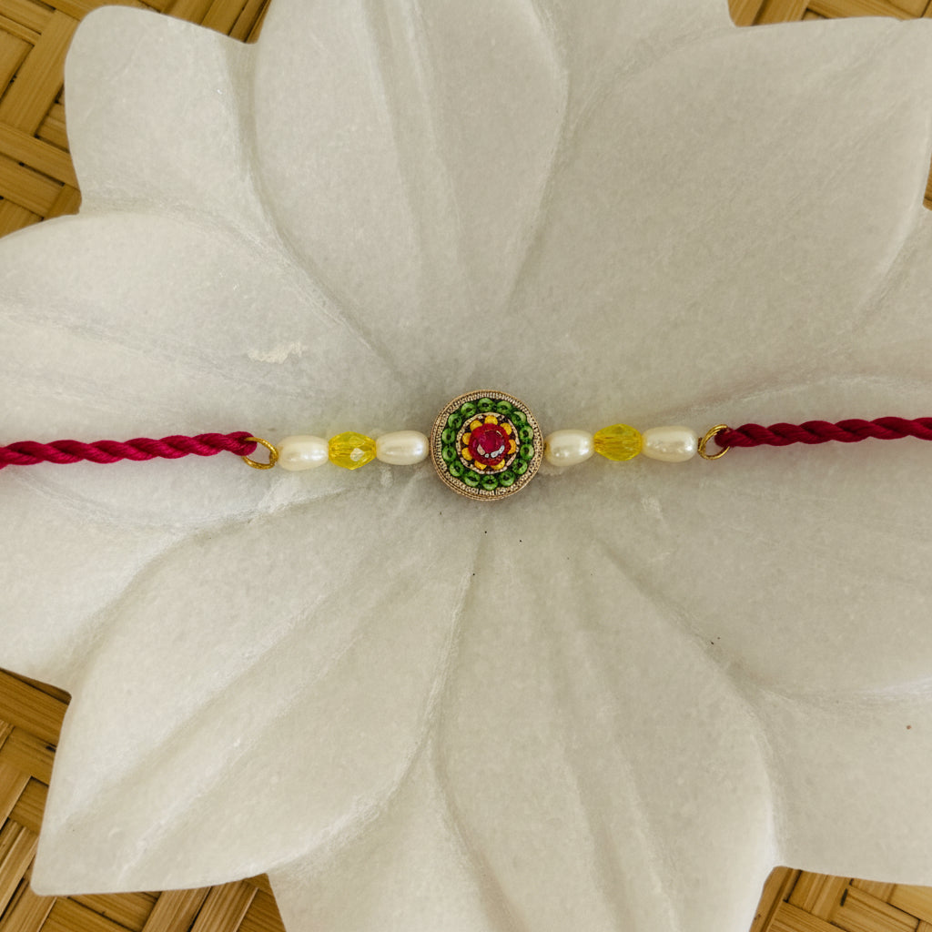A traditional Indian Rakhi bracelet with a red cord, white beads, yellow accents, and a central green and red paisley charm, displayed on a white lotus-shaped stand over a woven bamboo mat.