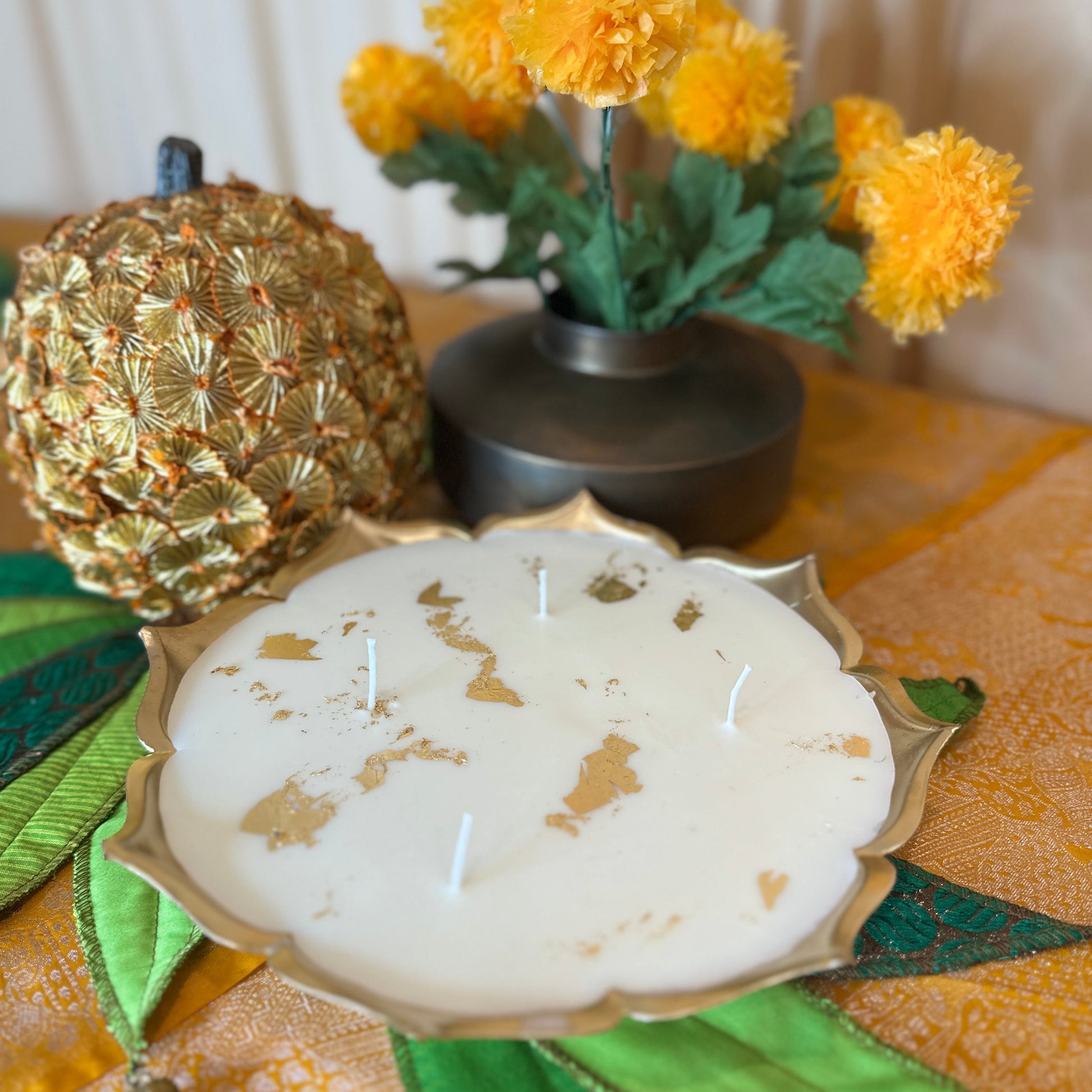 Decorative candle with gold leaf design on a table with a gold pumpkin and yellow flowers.